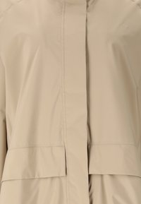 Beige vedenpitävä takki edestä, jossa peitetty vetoketju ja läppätaskut, esittäen sileää kangasta ja siistejä ompelusaumoja.