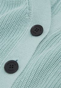 Ljusblå stickad tröja med ribbat mönster och två svarta knappar med präglad text. Närbild av tyg och knappdetalj.