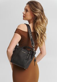 Sac seau en cuir noir avec une surface texturée, des accents dorés et une bandoulière ajustable. Il présente un haut froncé et une marque sur le devant.