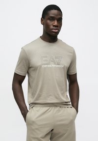 Άνδρας που φοράει μπεζ t-shirt EA7 Emporio Armani και ταιριαστά σορτς, στέκεται με τα χέρια στις τσέπες μπροστά σε απλό φόντο.