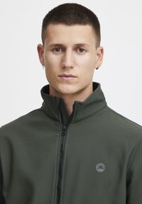 Dark green zip-up jacket made from flexible material. Features a high collar, black zipper, and a round black logo on the left chest.