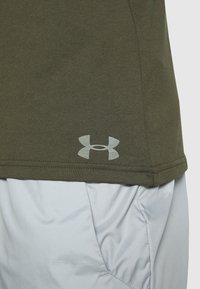 Olive green cotton blending shirt with a curved hem, featuring a light gray Under Armour logo along the bottom right corner.