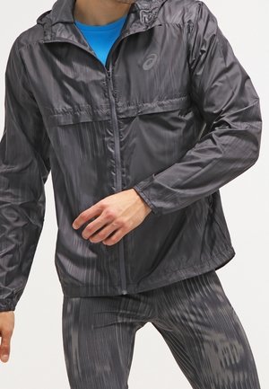 Mann trägt schwarzen Kapuzenzipper mit Muster und passende Sporthose mit blauem Shirt darunter, in Bewegung vor einfarbigem Hintergrund.