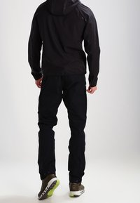 Veste imperméable noire à capuche au fini lisse, accompagnée de pantalons noirs et de chaussures marron avec des semelles vertes.