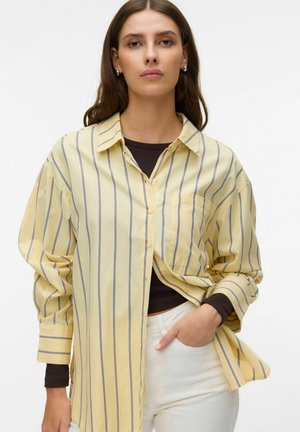 Vrouw draagt een loszittend geel overhemd met verticale zwarte strepen, een zwart shirt met lange mouwen eronder en witte broek, met één hand in haar zak.