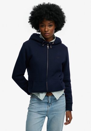 Kvinde med krøllet hår iført en marineblå hættejakke med lynlås over en stribet skjorte og lyseblå jeans, står med den ene hånd bag ryggen.