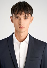 Granatowa marynarka z wyciętym klapem, pojedynczą kieszenią na piersi oraz białą, zapinaną na guziki koszulą pod spodem, charakteryzująca się gładką teksturą i dopasowanym krojem.