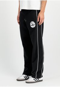 Schwarze Sweatpants mit weißen Seitenstreifen und dem Logo 'ALPHA INDUSTRIES'. Hergestellt aus weichem Stoff, mit elastischem Bund und Kordelzug.