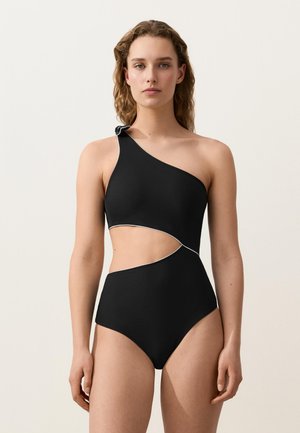 Kvinde med bølget hår iført en sort badedragt med ét skulderstrop og asymmetrisk udskæring med hvide kanter, stående mod en ensfarvet baggrund.