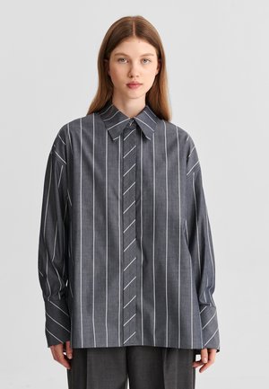 Femme aux cheveux lisses châtain clair portant une chemise ample gris foncé à rayures blanches verticales et col large, debout devant un fond uni.