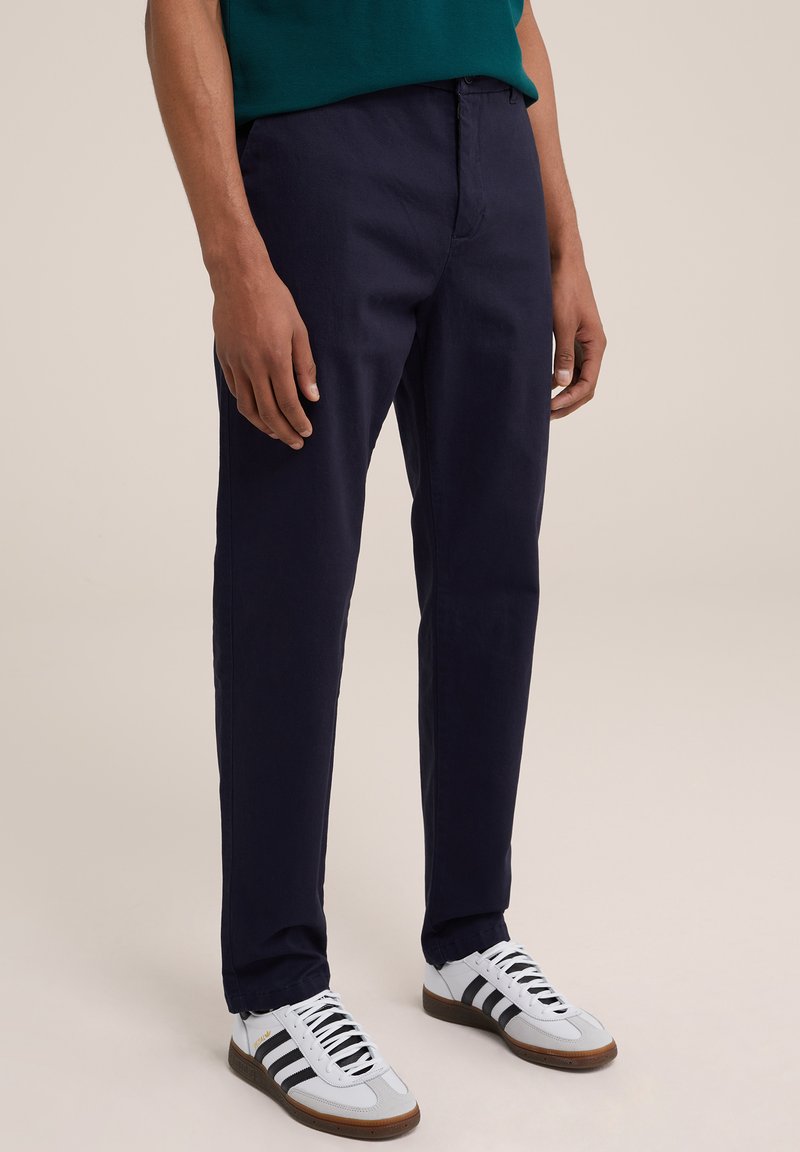 Pantaloni chino de culoare bleumarin, cu un design drept, având închidere cu nasture și bucle pentru curea, asortați cu adidași albi cu dungi negre.
