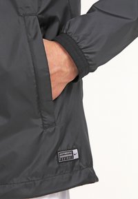 Chaqueta de nylon negra con puños acanalados y bolsillo lateral. Presenta una etiqueta que dice "FÚTBOL AUTÉNTICO" cerca del dobladillo. Textura suave.