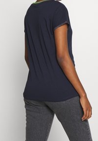 Námořnická modrá tričko s krátkým rukávem, s kulatým výstřihem a kontrastním zeleným lemem. Měkký bavlněný materiál s uvolněným střihem.