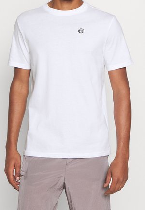 Man wearing plain white short-sleeve t-shirt with small black circular logo on left chest, paired with light gray textured pants.