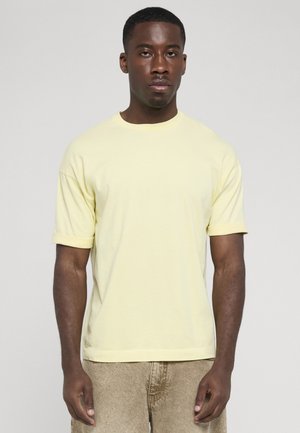Unge sort mand står iført en lys gul t-shirt og beige bukser mod en ensfarvet lysegrå baggrund.