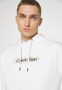 Felpa bianca in cotone con cappuccio regolabile, logo metallic oro e nero di Calvin Klein sul davanti e una trama morbida.