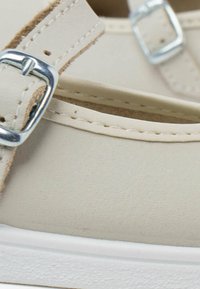 Zapato de cuero beige con una correa y hebilla plateada; presenta una textura suave y una suela de goma blanca, destacando la costura a lo largo de los bordes.
