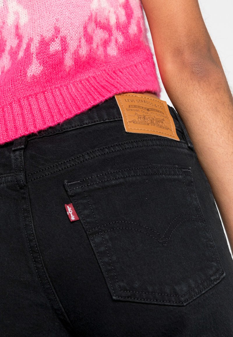Jeans en denim noir avec un patch logo en cuir, des détails en tab rouge et des surpiqûres contrastantes. Les jeans sont ajustés, présentant une surface texturée.