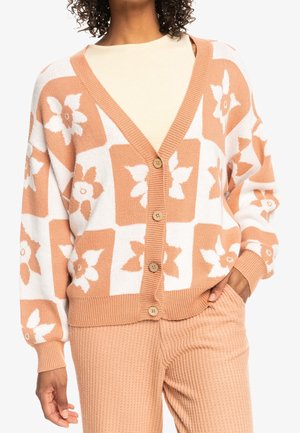 Mujer con un cárdigan de botones con patrón floral en tonos melocotón y blanco, pantalones de punto melocotón con canalé, con la mano en el bolsillo, sobre un fondo blanco.