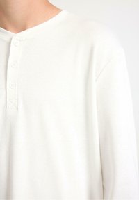 Langdurig mouw witte henleyshirt in geribbelde textuur, met een V-vormige halslijn en drie knopen aan de voorkant, afgewerkt met een gladde stof.