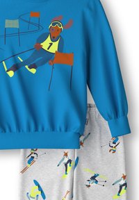 Een blauwe sweatshirt met een hond in skikleding, gecombineerd met grijze broek versierd met verschillende illustraties van skiën en snowboarden.