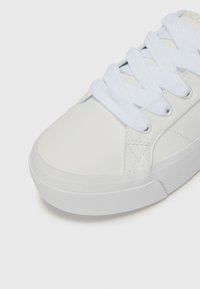 Hvit lær sneaker med en glatt tekstur, som har flate hvite lisser, en rund tå og en gummisåle med teksturert grep.