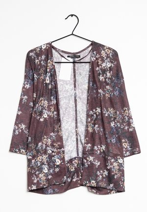 Gilet ouvert marron à fleurs avec des manches longues suspendu sur un cintre noir contre un fond blanc.