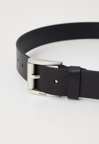 Svart lærbelte med en sølvfarget metallspenne. Spennen har et flatt design, og beltet har jevnt fordelte hull for justering.