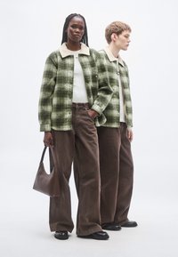 Groene geruite shacket met een crème sherpa-kraag, gecombineerd met bruine wijde broekspijpen en een bruine handtas. Soepele stoftextuur.