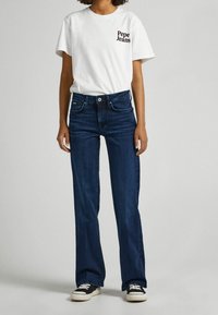 Camiseta blanca con el logo "Pepe Jeans", vaqueros acampanados oscuros y zapatillas negras con suelas blancas. Conjunto casual, diseño y corte simples.