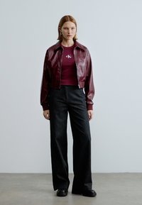 Bordeauxrote Lederjacke, kurz geschnitten, über einem dunkelroten T-Shirt mit Logo, kombiniert mit weitem schwarzen Denim-Hose und schwarzem Schuhwerk. Glatte Textur.