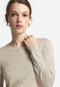 Beige långärmad tröja gjord av lätt material, med rund halsringning och åtsittande ärmslut. Modellen har silvriga hoops-örhängen.
