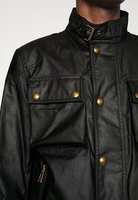 Černá bunda s lesklou texturou, zdobená zlatými kovovými detaily, vysokým límcem a několika kapsami na přední straně.