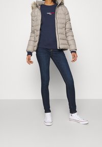 Person wearing beige puffer jacket, navy Tommy Jeans sweatshirt, dark blue skinny jeans, and white high-top Converse sneakers.