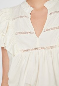 Blusa crema con scollatura arricciata, caratterizzata da inserti in pizzo verticali e maniche corte a palloncino. Tessuto morbido con un design fluido.