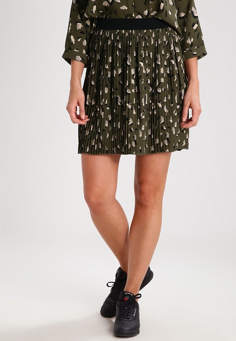 Femme portant une jupe plissée vert olive avec un imprimé abstrait beige et noir, associée à des baskets noires, debout les jambes croisées.