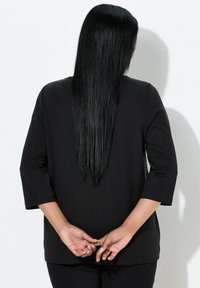Czarna bluzka z długim rękawem, okrągłym dekoltem i rękawami o długości 3/4. Gładka faktura, luźny krój; modelka stoi z włosami opadającymi na plecy.