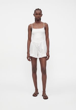 Femme portant une combinaison blanche sans manches avec un short à volants et des tongs noires, debout devant un fond clair uni.