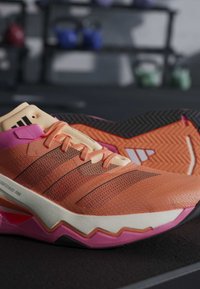 Chaussure de sport orange et rose avec des rayures noires et une semelle texturée posée sur une surface noire avec du matériel de gym flou en arrière-plan.