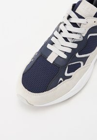 Navy blue sneaker with mesh and suede materials, white rubber sole, prominent lace-up design, and curved accents along the sides.