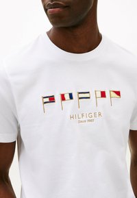 Maglietta bianca in cotone con bandiere colorate ricamate e la scritta "HILFIGER Since 1985" in oro sotto il design. Maniche corte e scollo rotondo.