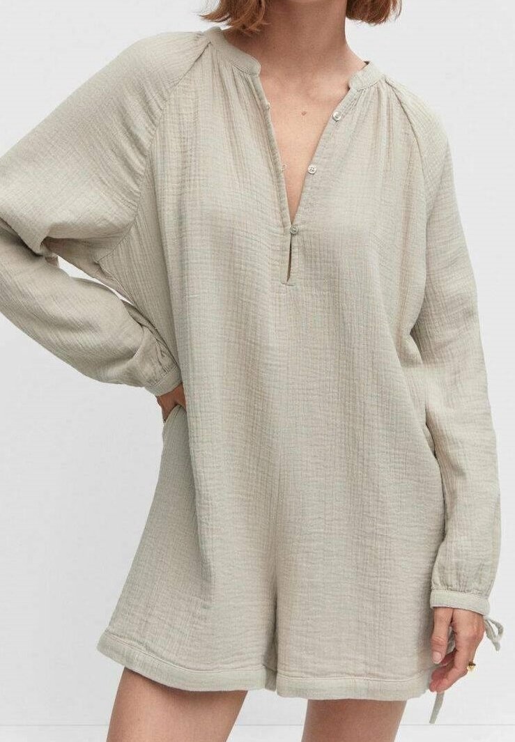 Femme portant une combinaison ample beige à manches longues, texturée, avec un col en V et des détails de boutons, debout avec une main sur la hanche.