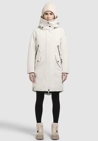Lange, witte parka met hoge kraag en capuchon, voorzien van een ritssluiting aan de voorkant, twee zijzakken en zwarte accenten. Gekenmerkt door zwarte leggings en beige laarzen.