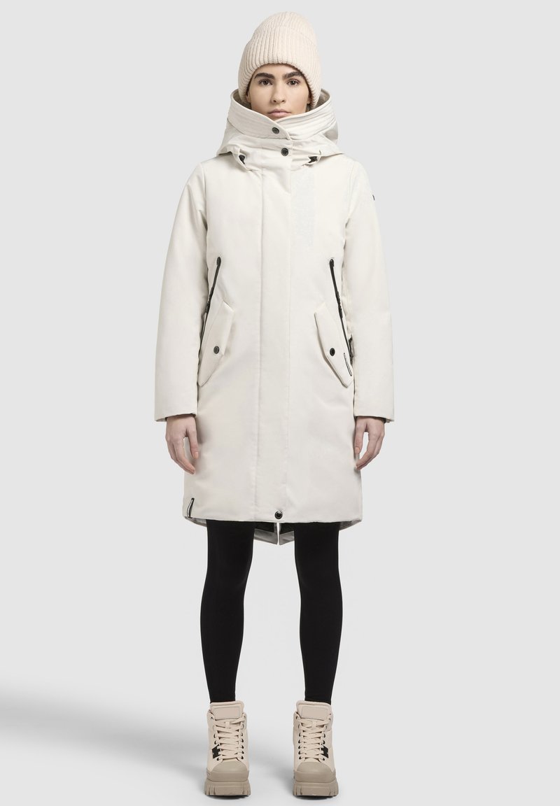 Lange, witte parka met hoge kraag en capuchon, voorzien van een ritssluiting aan de voorkant, twee zijzakken en zwarte accenten. Gekenmerkt door zwarte leggings en beige laarzen.