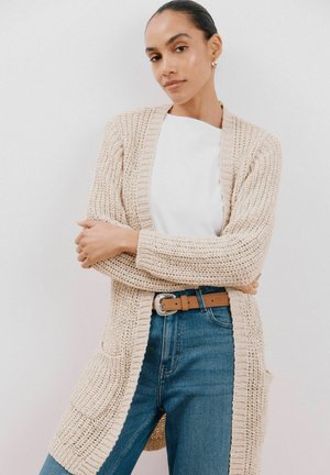 Hellbeige gestrickte Strickjacke mit offenem Vorderteil, langen Ärmeln und zwei Vordertaschen. Kombiniert mit einem weißen Oberteil und blauen Jeans, die einen braunen Gürtel tragen.