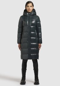 Ilga, tumši zaļa pūkainā mēteli ar spīdīgu apdari, ar kapuci un ar rāvējslēdzēja kabatām. Iezīmē taisnstūra siltumizolācijas rakstu un taisnu griezumu.