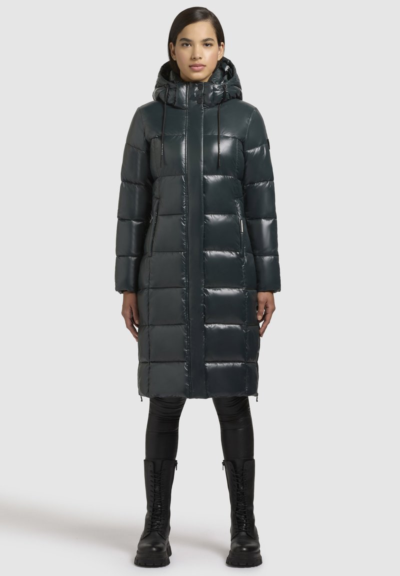 Ilga, tumši zaļa pūkainā mēteli ar spīdīgu apdari, ar kapuci un ar rāvējslēdzēja kabatām. Iezīmē taisnstūra siltumizolācijas rakstu un taisnu griezumu.