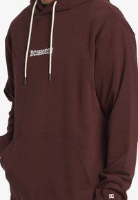 Burgundská mikina s kapucí vyrobená z měkké tkaniny. Obsahuje přední kapsu, bílé šňůrky a vyšitý logo "DCSHOECO".