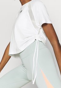 Top blanco corto con lazos laterales y paneles transparentes, combinado con leggings de talle alto en verde menta claro con una franja de acento durazno.