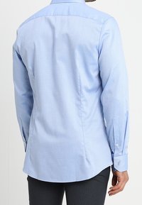 Camicia azzurra a maniche lunghe con polsini a bottoni, lieve texture e orlo curvo. Presenta un colletto standard e un dettaglio sul giromanica posteriore.